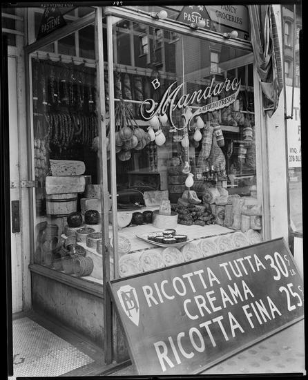 Black and white image of Storefront in the Italian quarter of Chicago, with a sign on the window saying 'Mandaro' and sidewalk sign advertising Ricotta Tutta, Crema, Ricotta Fina