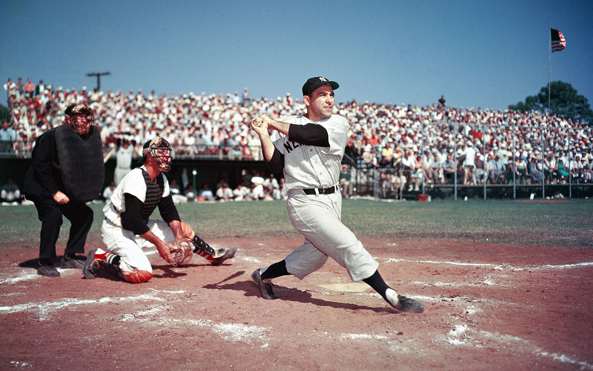 Old photo of Yogi hitting swinging at the plate in Yankees uniform.