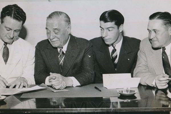 Three men in suits overlooking Lou Gehrig signing a contract