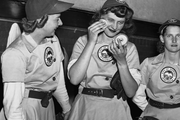 Three female baseball players from the Kenosha Comets in the dugout, with one applying makeup from a compact