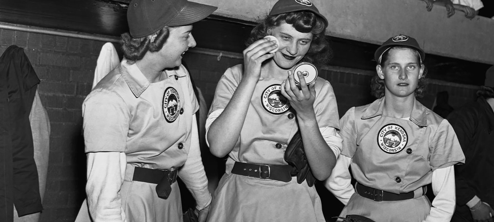 Three female baseball players from the Kenosha Comets in the dugout, with one applying makeup from a compact