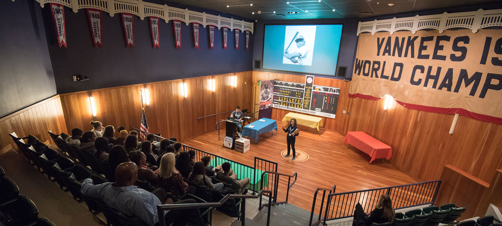 A guide talking to an audience in the Museum's auditorium with an image of Jackie Robinson on the screen behind her