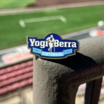 Image of a Pin sitting on a railing overlooking a baseball field. The Pin reads Yogi Berra with the subtitle 1925:2025 Centennial