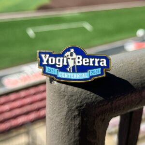 Image of a Pin sitting on a railing overlooking a baseball field. The Pin reads Yogi Berra with the subtitle 1925:2025 Centennial